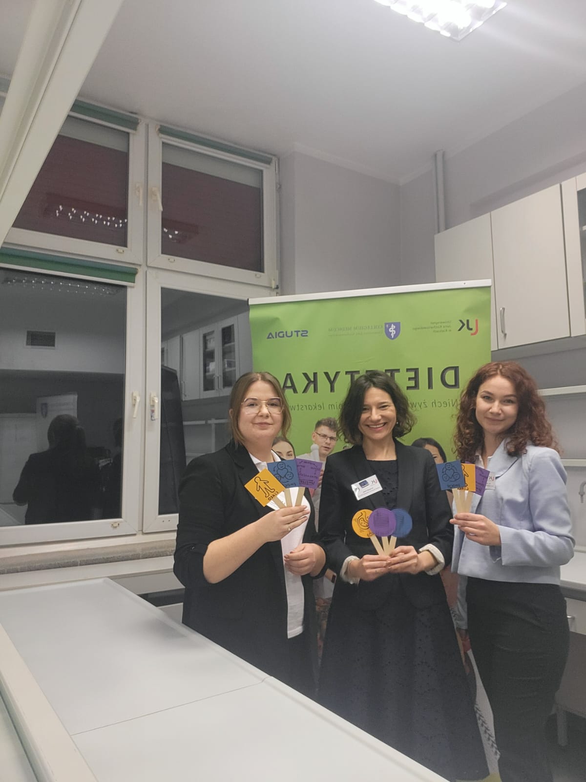 Obraz przedstawia wnętrze, w którym osoby stoją przed jasnym banerem i trzymają kolorowe papierowe tabliczki. Pomieszczenie jest jasne, z neutralnymi ścianami i oświetleniem sufitowym. W tle widoczne jest okno z zasłoniętymi roletami oraz przeszklona szafka. Całość sugeruje wydarzenie o charakterze prezentacyjnym lub edukacyjnym.