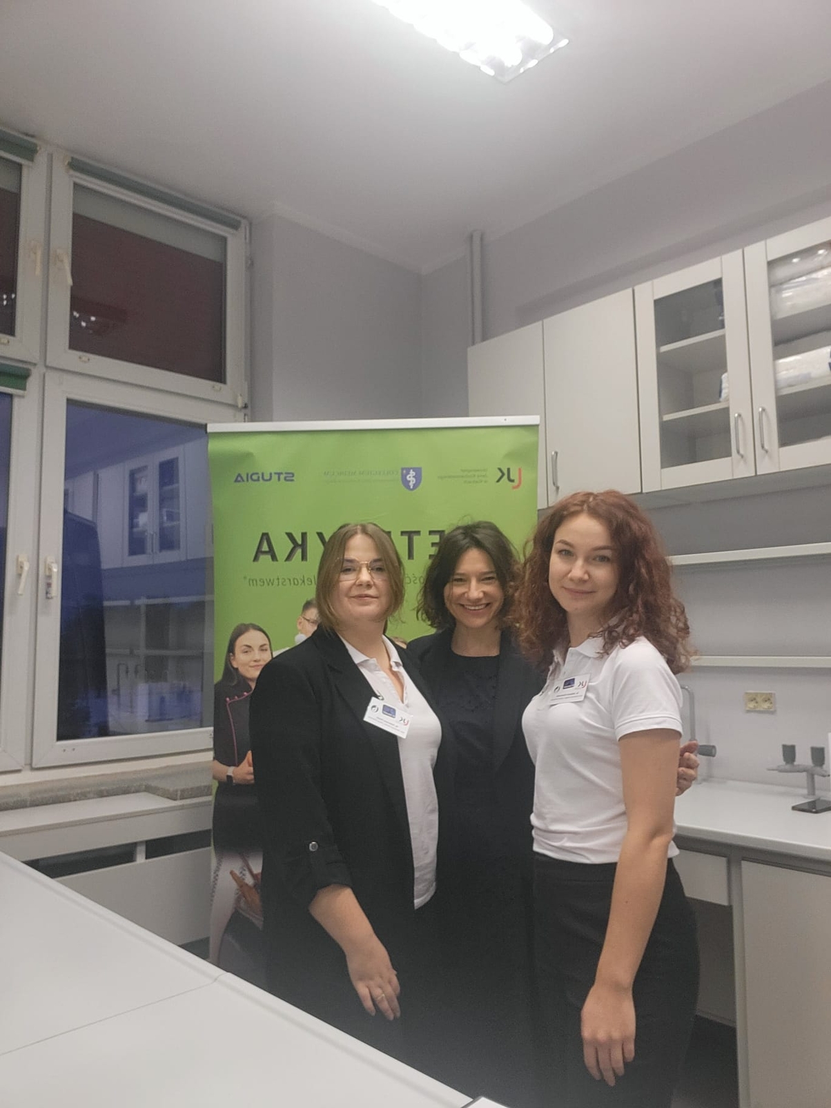 Obraz przedstawia osoby stojące w sali laboratoryjnej lub edukacyjnej, na tle zielonego baneru z napisami i logotypami. W pomieszczeniu widoczne są szafki oraz blaty charakterystyczne dla takiej przestrzeni. Nad głowami znajduje się oświetlenie sufitowe, a z boku widać duże okna odbijające wnętrze sali. Scena sugeruje wydarzenie o charakterze edukacyjnym lub prezentacyjnym.