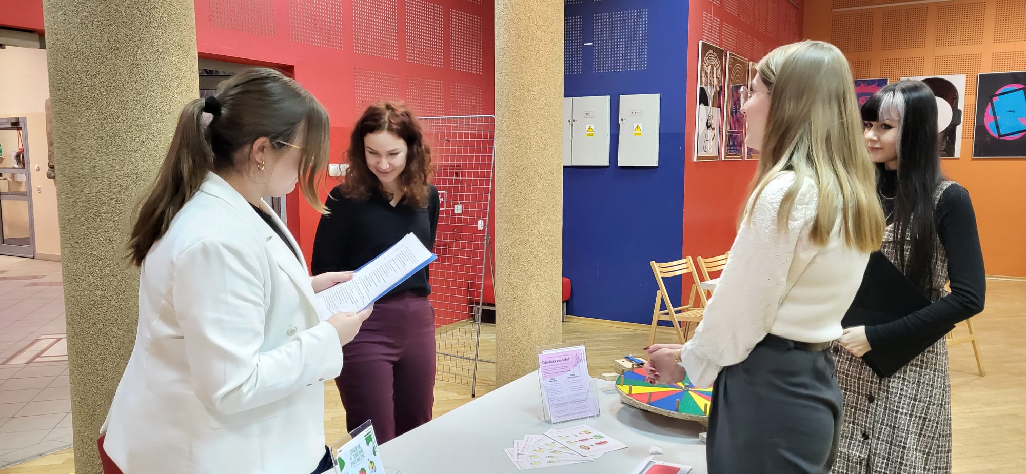 Obraz przedstawia spotkanie w pomieszczeniu, w którym osoby stoją przy stole z materiałami informacyjnymi. Jedna z osób odczytuje treść z dokumentu, a pozostałe słuchają i obserwują. Na stole znajdują się ulotki oraz element edukacyjny o formie gry. W tle widoczne są plakaty na ścianach, ustawione krzesła oraz elementy infrastruktury technicznej. Scena sugeruje wydarzenie informacyjne lub edukacyjne.