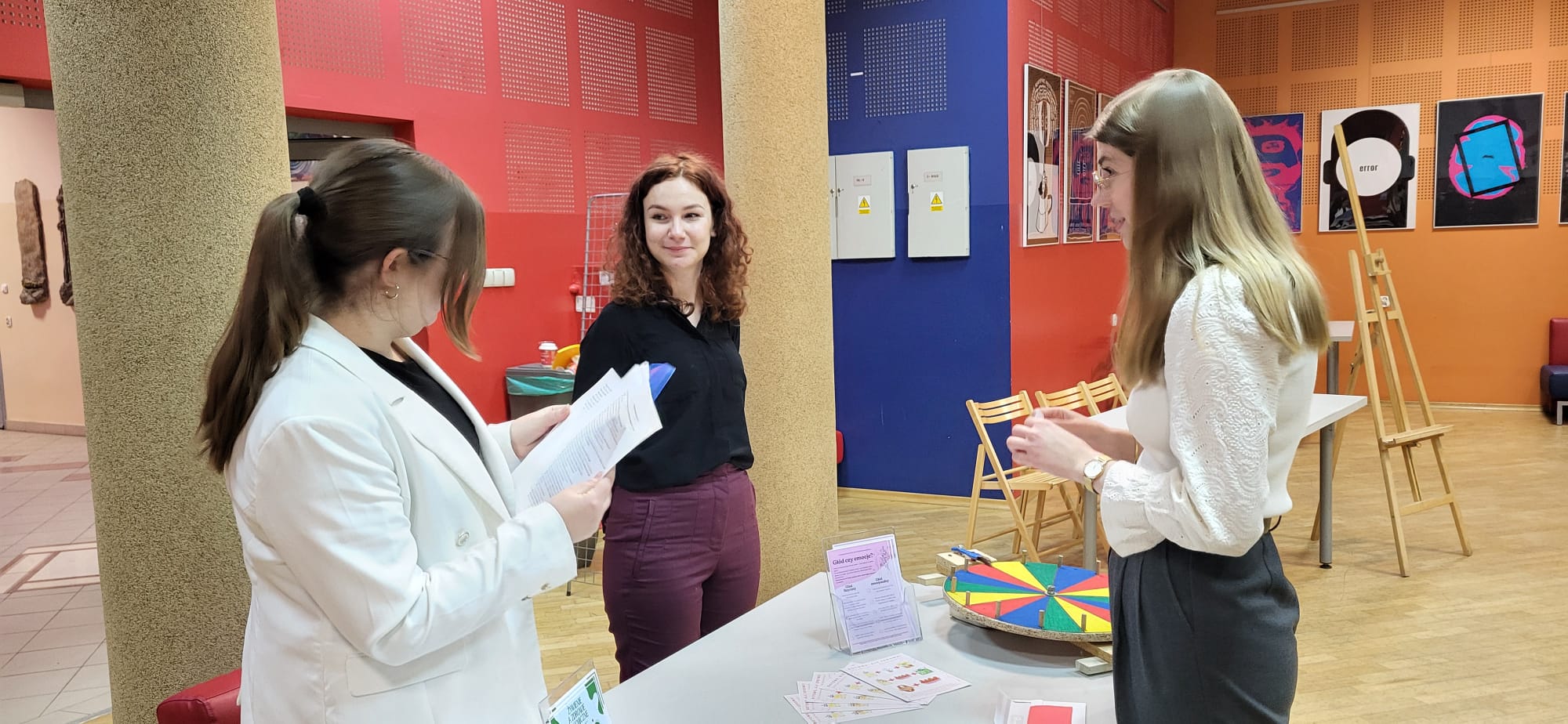 Obraz przedstawia osoby stojące wokół stołu w jasnym pomieszczeniu. Jedna z nich trzyma dokumenty i na nich się skupia, inna obserwuje sytuację z zainteresowaniem, a kolejna korzysta z kolorowego koła edukacyjnego znajdującego się na stole. W tle widoczne są ściany z oprawionymi pracami, elementy architektoniczne oraz część zaplecza z drzwiami i krzesłami. Scena sugeruje aktywność o charakterze edukacyjnym lub informacyjnym.