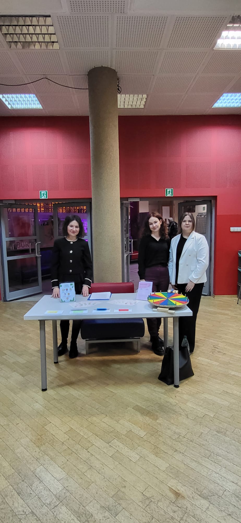 Obraz przedstawia wnętrze sali z parkietem i czerwonymi ścianami. Za stołem z uporządkowanymi, kolorowymi materiałami oraz elementem edukacyjnym w formie obrotowego koła stoją uczestnicy wydarzenia. W tle widoczna jest kolumna, drzwi prowadzące do innej przestrzeni oraz oznaczenia ewakuacyjne. Całość sugeruje spotkanie lub aktywność o charakterze informacyjnym albo edukacyjnym.