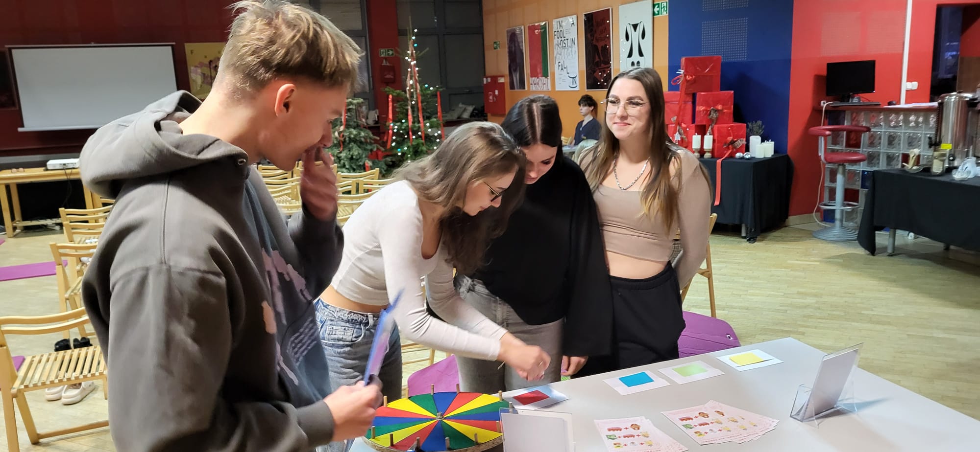Obraz przedstawia osoby zgromadzone wokół stołu w sali z drewnianą podłogą i dekorowanymi ścianami. Na stole znajdują się kolorowe materiały oraz element do gry lub ćwiczeń edukacyjnych. Uczestnicy są zaangażowani w aktywność. W tle widać krzesła, ekran projekcyjny, plakaty oraz dekoracje świąteczne na bocznym stoliku. Scena sugeruje warsztaty lub zajęcia edukacyjne.