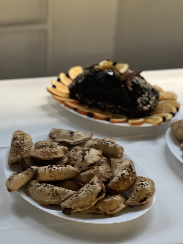 Na zdjęciu widać stół z dwoma daniami. Na pierwszym planie znajduje się talerz z pieczonymi, półksiężycowatymi wypiekami posypanymi czarnymi ziarnami. W tle jest deser o podłużnym kształcie z polewą, udekorowany kawałkami pomarańczy. Jasne tło podkreśla wygląd potraw.