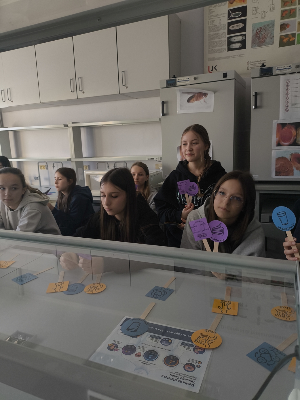 Uczniowie w sali lekcyjnej trzymają pomoce dydaktyczne na patyczkach. W otoczeniu widoczny jest sprzęt laboratoryjny oraz plakaty edukacyjne, co wskazuje na zajęcia o charakterze naukowym.
