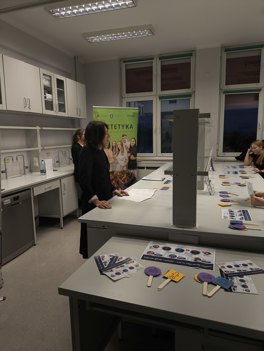 Sala lekcyjna, w której uczestnicy stoją wokół stołu z materiałami edukacyjnymi oraz tabliczkami z symbolami. Przy ścianie widoczny jest plakat, co wskazuje na zajęcia o charakterze dydaktycznym.