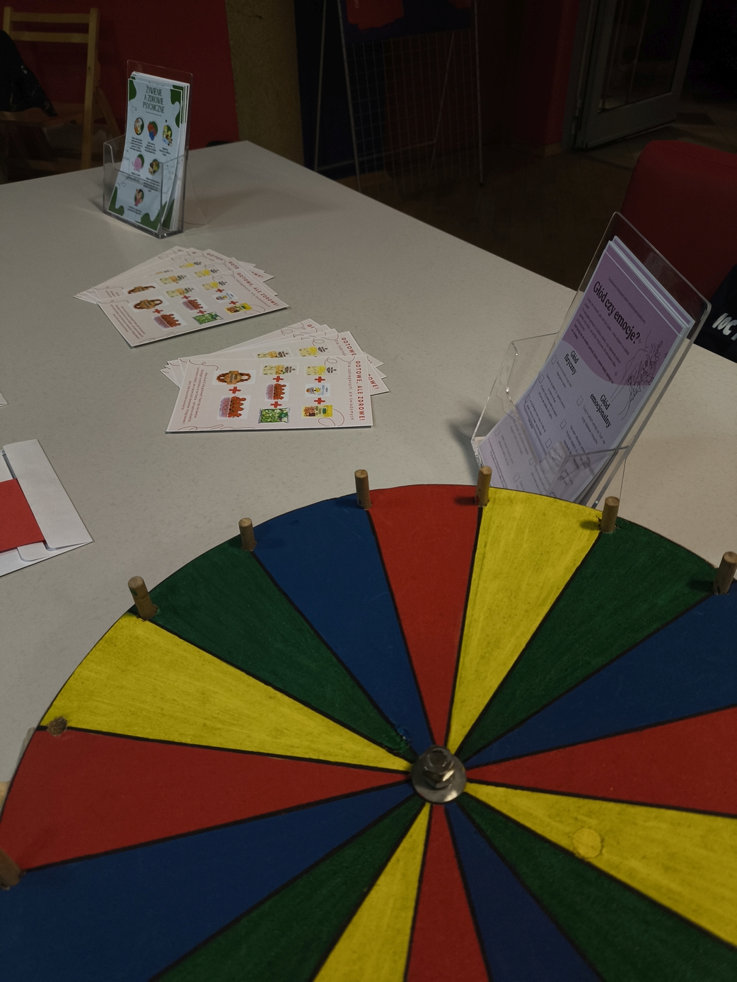 Obraz przedstawia stół z kolorowym drewnianym kołem obrotowym podzielonym na barwne segmenty. Na jego krawędzi znajdują się małe drewniane elementy. W tle widoczne są materiały edukacyjne lub informacyjne, takie jak kartki z grafikami oraz stojaki z broszurami. Całość znajduje się w pomieszczeniu wewnętrznym, przypominającym przestrzeń warsztatową lub wystawową.