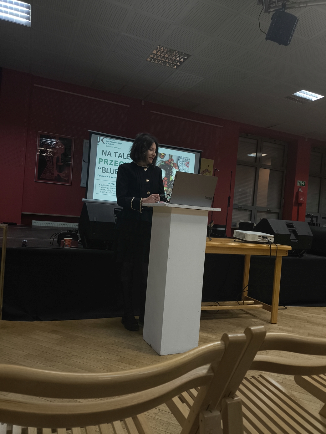 Obraz przedstawia osobę prowadzącą prezentację przy białej mównicy w sali z drewnianą podłogą. W tle widoczna jest ściana oraz ekran z wyświetloną prezentacją. Obok znajduje się stół ze sprzętem technicznym i głośnik. Wnętrze ma oświetlenie sufitowe, a na pierwszym planie częściowo widać krzesła. Scena sugeruje wykład lub wystąpienie.