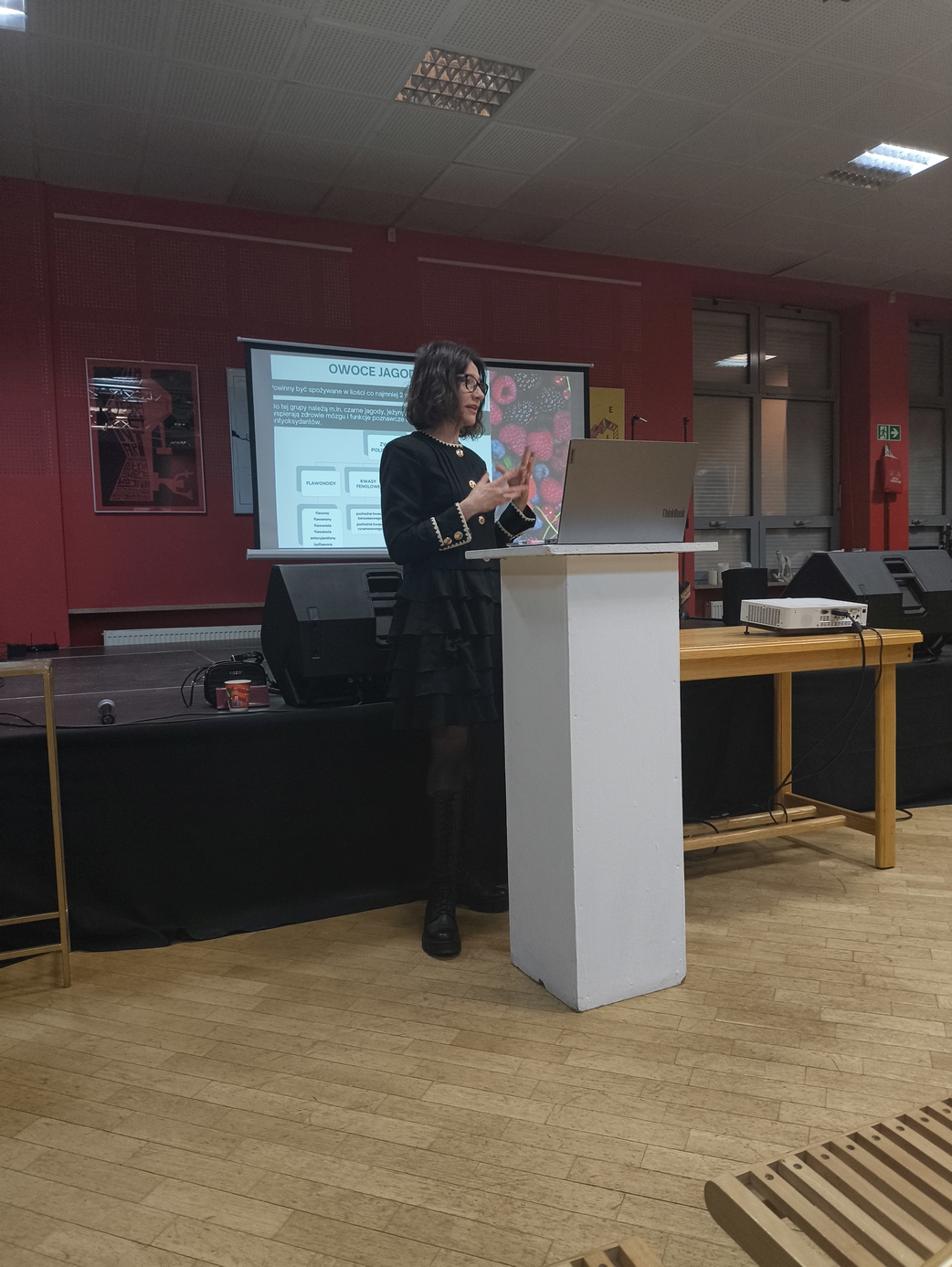 Obraz przedstawia osobę prowadzącą prezentację przy białej mównicy w sali z drewnianą podłogą. W tle widoczna jest ściana oraz ekran z wyświetloną prezentacją. Obok znajduje się stół ze sprzętem technicznym i głośnik. Wnętrze ma oświetlenie sufitowe, a na pierwszym planie częściowo widać krzesła. Scena sugeruje wykład lub wystąpienie.