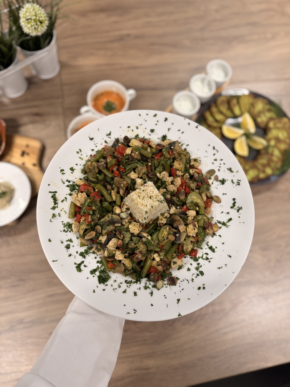 Na zdjęciu widać biały talerz z kolorowym stir-fry z warzyw (fasolka, papryka, cukinia, pieczarki) i kawałkami tofu, podanymi z porcją ryżu i posypanymi natką pietruszki. W tle są sosy, ogórki z cytryną, zupa oraz dekoracyjne kwiaty.