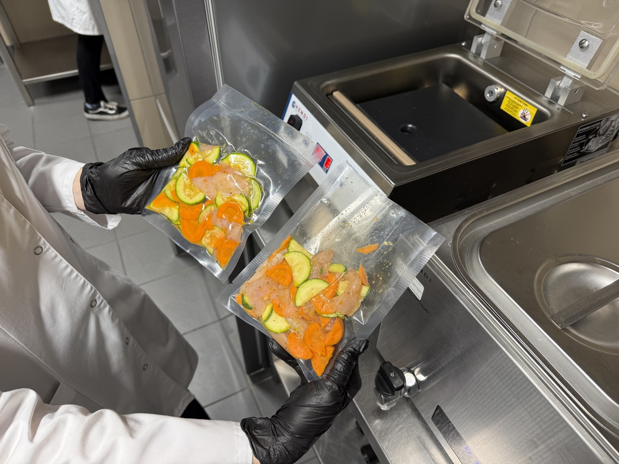 Na zdjęciu widać osobę w białym fartuchu i czarnych rękawicach, trzymającą dwie zapakowane próżniowo torby z warzywami (marchew, cukinia) i surowym kurczakiem. Scena rozgrywa się w kuchni obok zgrzewarki próżniowej, a w tle widoczna jest druga osoba.