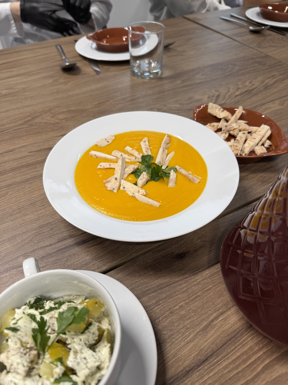 Na zdjęciu widać stół z posiłkiem: miskę pomarańczowej zupy z grzankami i ziołem oraz małą miseczkę z kremową sałatką. Obok stoi miseczka z dodatkowymi grzankami, a w tle widać zastawę stołową, dzbanek i częściowo widoczne osoby przy stole.