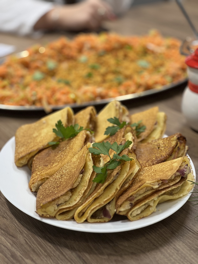Na zdjęciu widać naleśniki złożone w trójkąty, ułożone na białym talerzu na drewnianym stole. Są udekorowane świeżą natką pietruszki. W tle znajduje się rozmyte danie z różnorodnych składników, a po prawej stronie częściowo widoczny jest niewielki pojemnik z czerwonym wieczkiem.