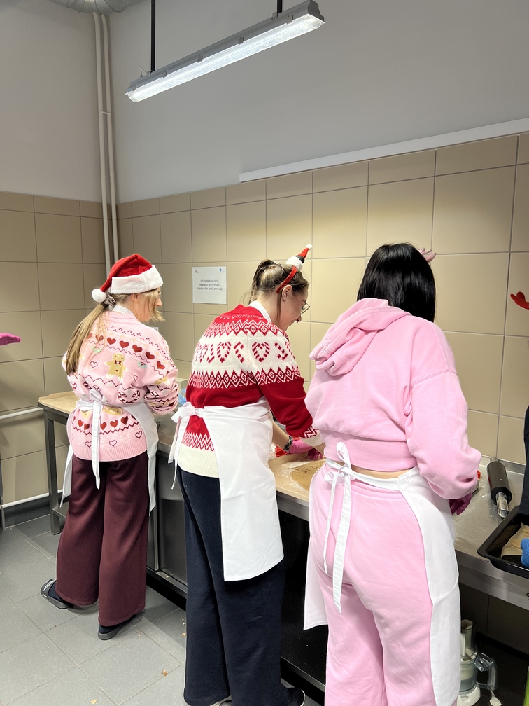 Na zdjęciu widać osoby w kuchni, które wspólnie przygotowują wypieki przy blacie. Pracują w jasnym, wyłożonym płytkami wnętrzu, oświetlonym lampą sufitową. Atmosfera sugeruje wspólne, świąteczne gotowanie.