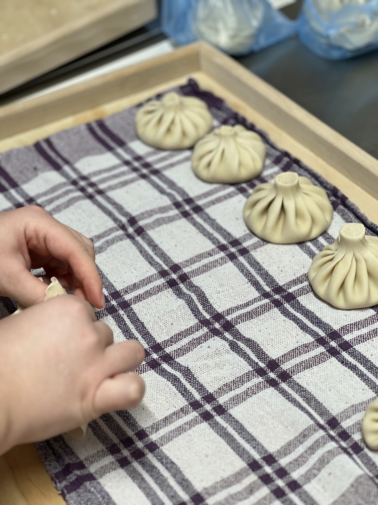 Na zdjęciu widać przygotowywanie pierogów na drewnianej tacy wyłożonej biało-fioletową ściereczką. Uformowane pierogi z charakterystycznym falowanym brzegiem są ułożone w rzędzie, a obok trwa formowanie kolejnego.