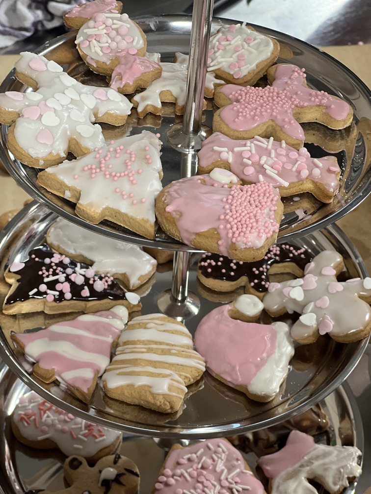 Na zdjęciu widać dwupoziomową paterę z dekorowanymi ciasteczkami o świątecznych kształtach. Ciasteczka są pokryte białym, różowym i czekoladowym lukrem oraz ozdobione kolorowymi posypkami. Całość ma słodki, świąteczny charakter.