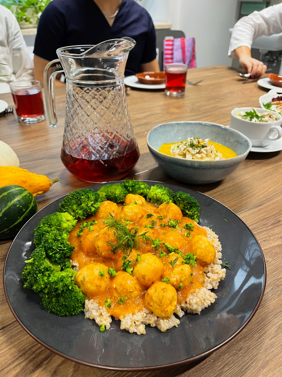 Na zdjęciu widać stół z posiłkiem. Na pierwszym planie jest talerz z małymi pierogami w pomarańczowym sosie, brokułem i brązowym ryżem, udekorowanymi ziołami. Obok stoi dzbanek z czerwonym napojem, a w tle widać osobę przy stole, inne dania i ozdobną dynię.