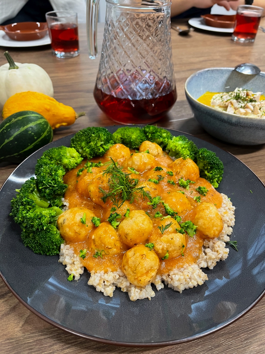 Na zdjęciu widać stół z różnymi potrawami i napojami. Na pierwszym planie jest czarny talerz z kulkami w pomarańczowym sosie, brokułem i jasną kaszą, udekorowany ziołami. W tle stoją dzbanek i szklanka z czerwonym napojem, ozdobne dynie oraz miska z innym daniem, co sugeruje wspólny posiłek.