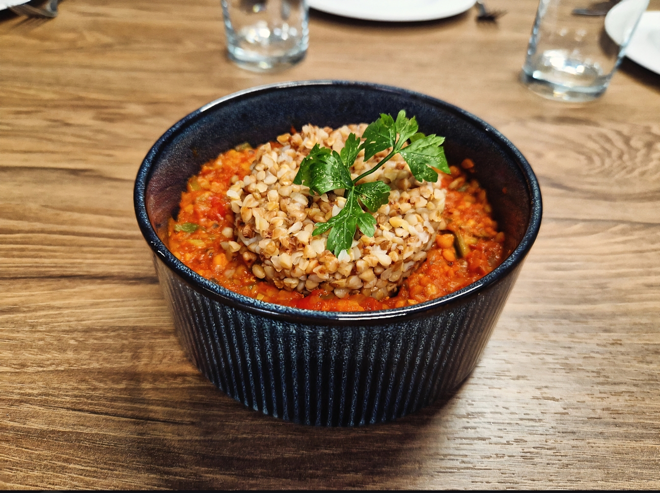 Na zdjęciu widać granatową, ceramiczną miskę z daniem z czerwonym sosem warzywnym, na którym znajduje się porcja kaszy, prawdopodobnie gryczanej. Całość udekorowana jest natką pietruszki i stoi na drewnianym stole, a w tle widać rozmyte elementy zastawy stołowej.