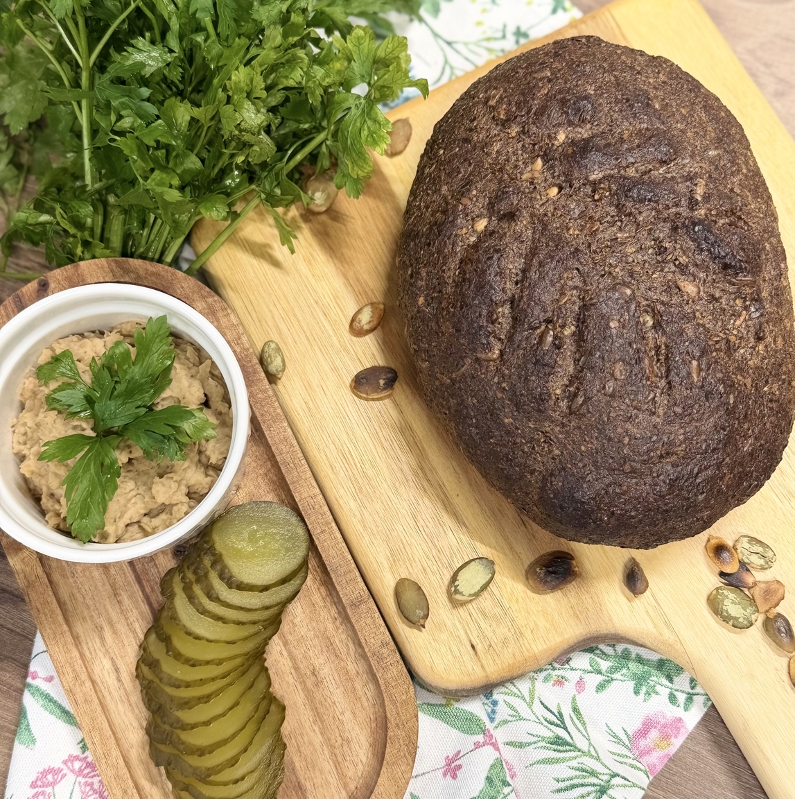 Na zdjęciu widać rustykalną aranżację jedzenia na drewnianej powierzchni. Po prawej leży bochenek ciemnego, ziarnistego chleba na desce do krojenia. Po lewej znajduje się tacka z ogórkami kiszonymi oraz miseczka z jasnym dipem udekorowanym natką pietruszki. Całość uzupełniają pestki dyni i kwiatowa tkanina w tle.