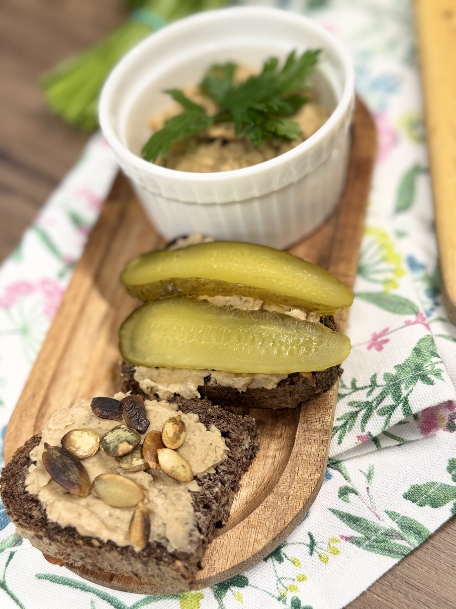 Na obrazie widać drewnianą deskę z dwiema kromkami ciemnego chleba żytniego posmarowanymi jasną pastą. Jedna kromka jest udekorowana plasterkami ogórka, druga pestkami, m.in. dyni. W tle znajduje się biała miseczka z pastą i natką pietruszki oraz delikatnie widoczna tkanina w kwiaty i seler.