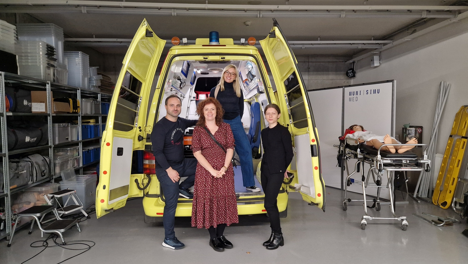 Na obrazie widać kilka osób stojących przy otwartym tyle żółtej karetki pogotowia. Drzwi ambulansu są otwarte, a wewnątrz znajduje się sprzęt medyczny. Obok karetki leży manekin medyczny umieszczony na noszach. W tle widoczne są regały z pojemnikami i materiałami oraz przegroda z napisem „NURSIMU MED”.