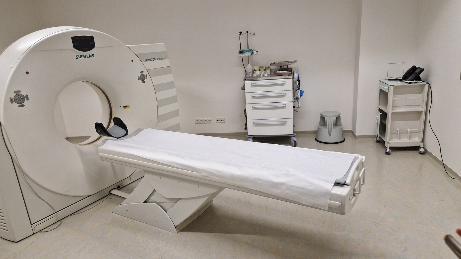 Na obrazie przedstawiono pracownię diagnostyki obrazowej z tomografem komputerowym. Urządzenie znajduje się po lewej stronie pomieszczenia i ma okrągły otwór oraz wysuwany stół do badania. W tle widoczny jest mobilny wózek z materiałami medycznymi oraz niewielki stołek. Po prawej stronie stoi drugi wózek na kółkach z telefonem podłączonym do ściany. Pomieszczenie jest jasne, czyste i ma charakter kliniczny.