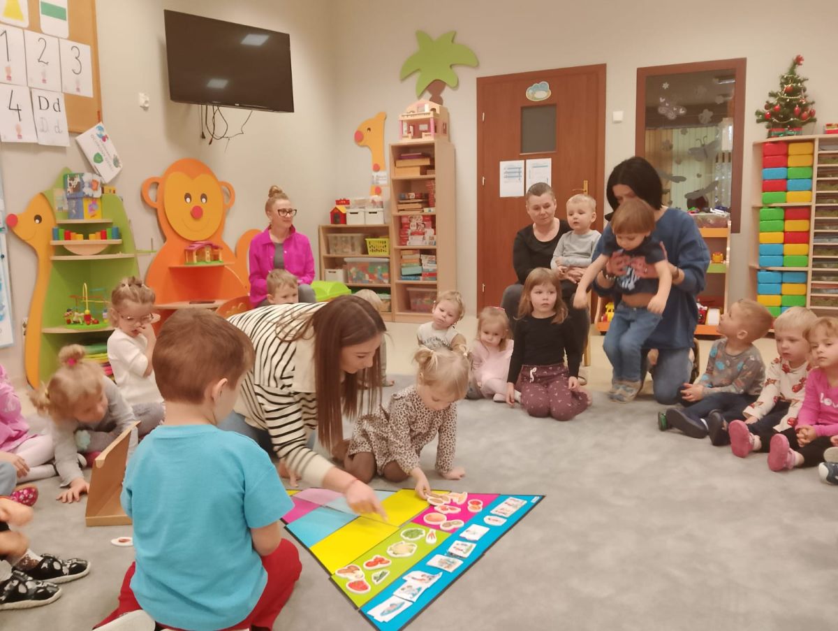 Obraz przedstawia salę przedszkolną, w której dzieci wraz z dorosłymi biorą udział we wspólnej aktywności. Dzieci siedzą na dywanie wokół kolorowej planszy edukacyjnej umieszczonej na podłodze. Dorosły pomaga im w wykonywaniu zadania. W tle widoczne są meble, półki z zabawkami i książkami, tablica z pracami oraz elementy wyposażenia sali, takie jak drzwi i telewizor. Dzieci są zaangażowane w obserwowanie i udział w zabawie.