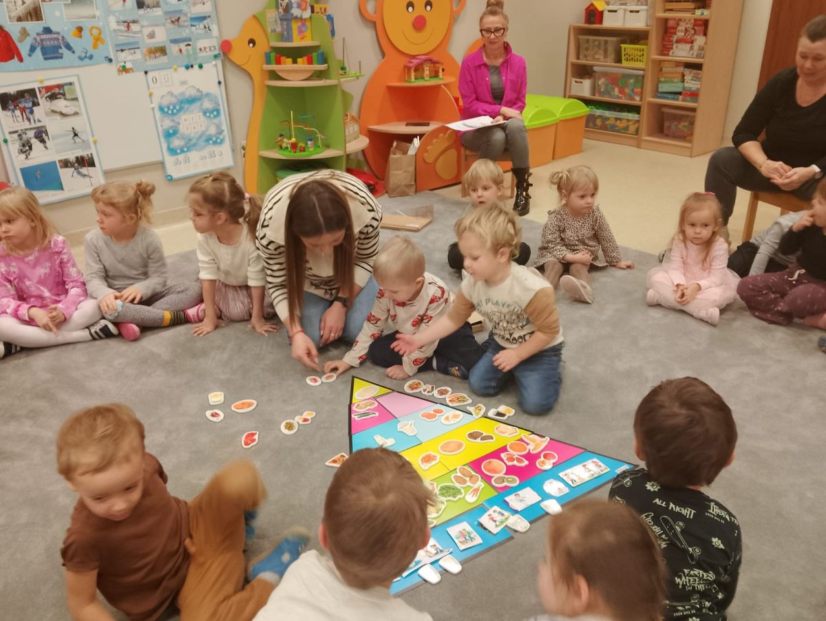 Obraz przedstawia salę przedszkolną, w której dzieci wraz z dorosłymi siedzą na dywanie i uczestniczą we wspólnej aktywności edukacyjnej. Dzieci skupione są wokół kolorowej planszy lub maty z ilustracjami, wykonując zadanie polegające na dopasowywaniu lub sortowaniu. Dorosły pomaga dzieciom w zabawie, a drugi obserwuje zajęcia. W tle widoczne są kolorowe meble, półki z materiałami edukacyjnymi i zabawkami oraz dekoracje na ścianach, tworzące przyjazne środowisko do nauki.