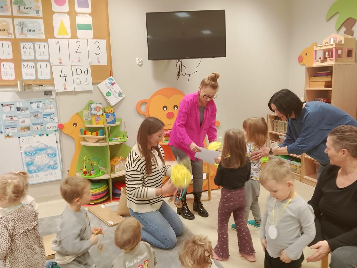 Obraz przedstawia salę przedszkolną, w której dzieci i dorośli biorą udział w zajęciach plastycznych. Na ścianach znajdują się plakaty edukacyjne z literami, cyframi i kształtami oraz telewizor. W sali widoczne są półki z zabawkami i pomocami dydaktycznymi. Dorośli pomagają dzieciom w pracy z materiałami plastycznymi. Całość tworzy kolorowe, przyjazne i edukacyjne otoczenie.