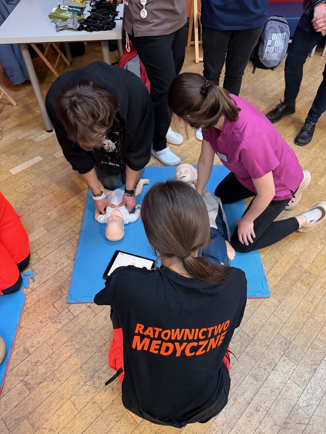 Zdjęcie przedstawia szkolenie z resuscytacji krążeniowo-oddechowej. Uczestnicy ćwiczą uciski klatki piersiowej na fantomie leżącym na macie. Jedna osoba nadzoruje przebieg szkolenia, a obok widoczny jest sprzęt medyczny.