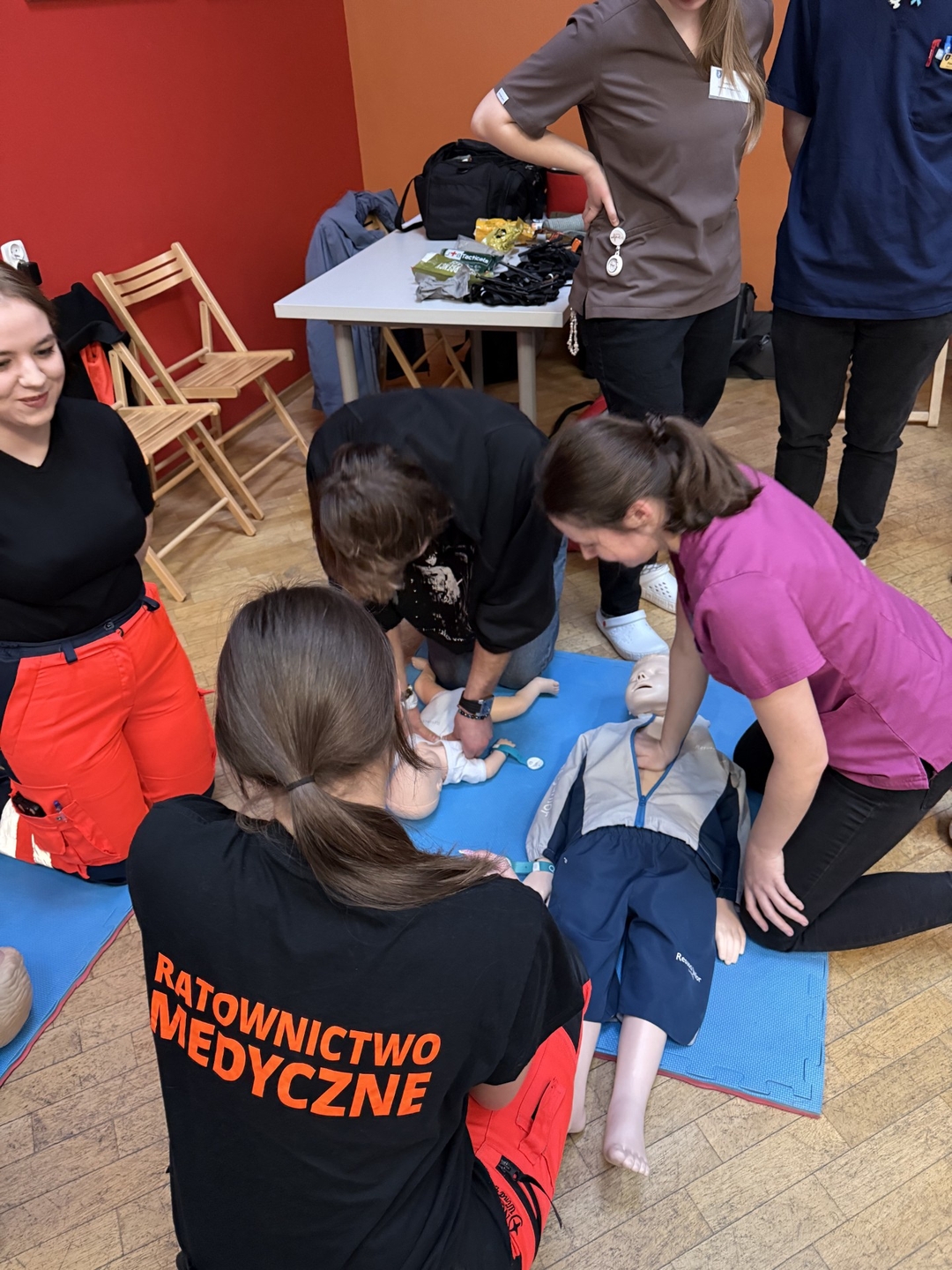 Grupa osób ćwiczy procedury medyczne podczas szkolenia z użyciem dwóch fantomów – dziecięcego i dla dorosłych. Zajęcia odbywają się w sali z kolorowymi ścianami, a fantomy znajdują się na macie. W tle widoczny jest stolik z różnymi przedmiotami oraz ustawione krzesła.