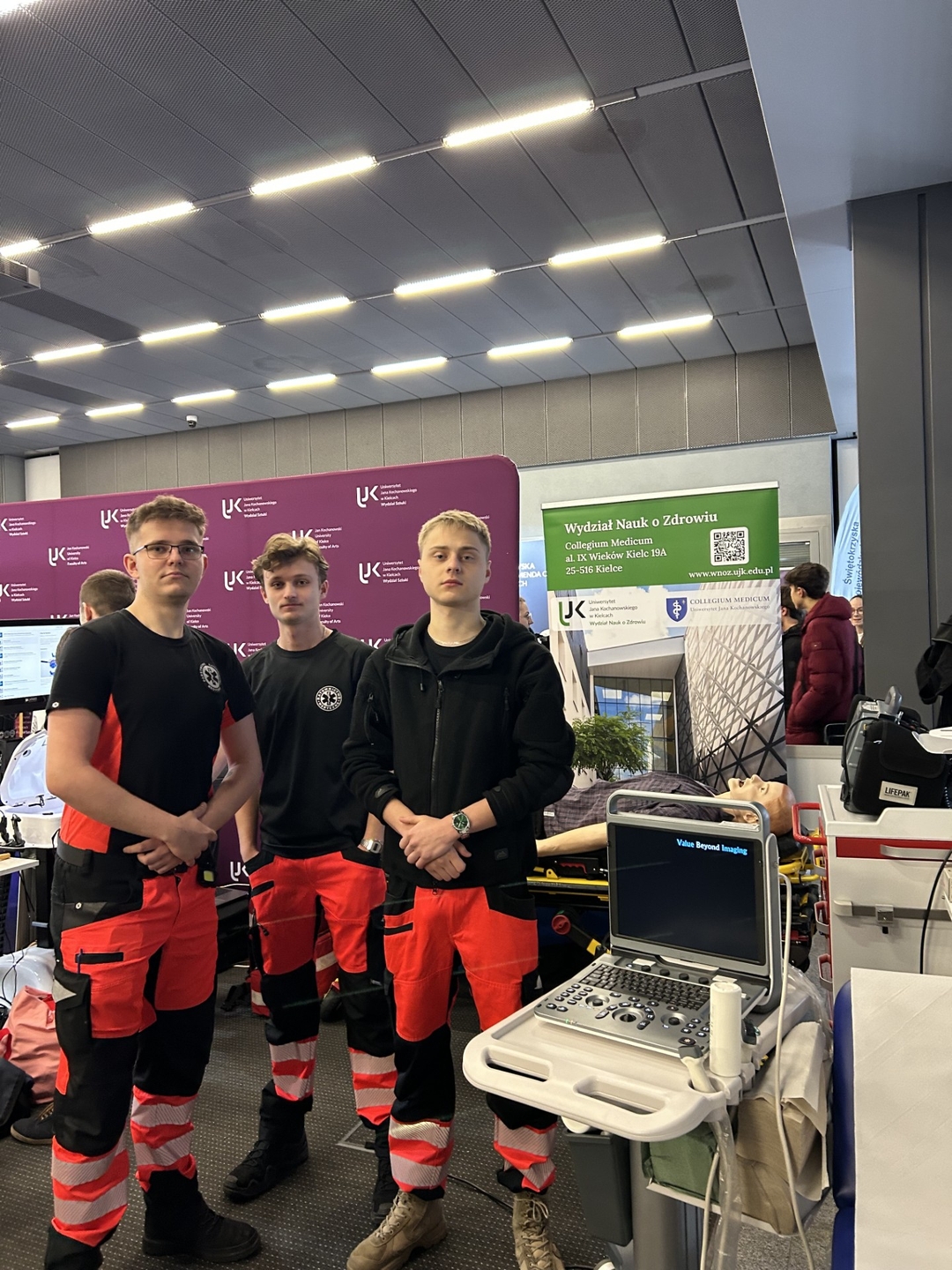 Obraz przedstawia trzy osoby stojące razem w dobrze oświetlonym wnętrzu. W tle znajduje się stoisko z różnym sprzętem medycznym, m.in. przenośnym aparatem USG. Widoczny jest także fantom leżący na noszach oraz sprzęt ratowniczy, w tym defibrylator. W przestrzeni znajdują się banery z tekstem i logo, co sugeruje, że scena ma miejsce w centrum wystawowym lub podczas targów.