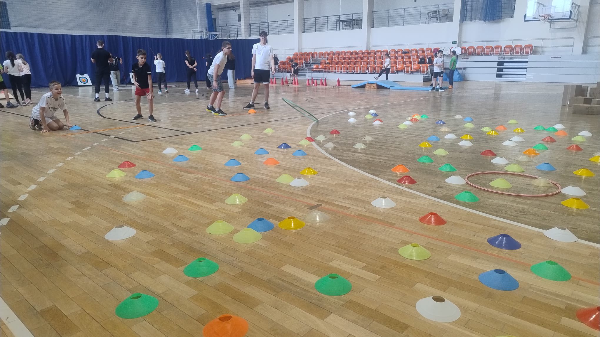 Zdjęcie przedstawia salę gimnastyczną z drewnianą podłogą. Na podłodze rozmieszczono kolorowe plastikowe pachołki (zielone, żółte, czerwone, niebieskie i białe), ułożone w określonym wzorze. Z przodu leży czerwone koło hula-hop położone na kilku pachołkach. W tle widać przygotowania do zabawy lub ćwiczeń. Przy ścianie znajduje się niebieska przegroda, dalej pomarańczowe siedzenia oraz schody. Sala jest przestronna, ma białe ściany i zamontowany kosz do koszykówki.