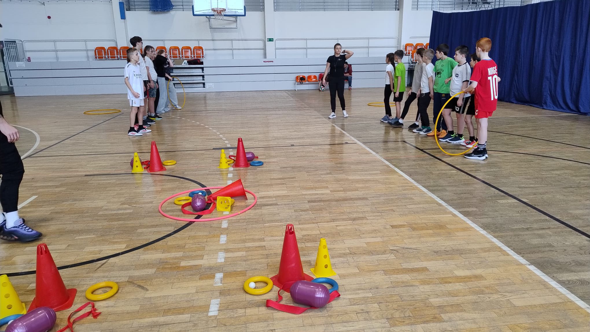 Zdjęcie przedstawia salę gimnastyczną z drewnianą podłogą. Dwie grupy dzieci stoją naprzeciwko siebie w rzędach. Na środku sali rozłożone są kolorowe przybory: czerwone i żółte pachołki, żółte obręcze, niebieskie krążki, czerwone wstążki oraz fioletowo-żółte woreczki. Pomiędzy grupami stoi osoba prowadząca zajęcia i udziela instrukcji. W tle widać ułożone pomarańczowe krzesła oraz kosz do koszykówki. Scena przedstawia lekcję wychowania fizycznego lub zajęcia sportowe.