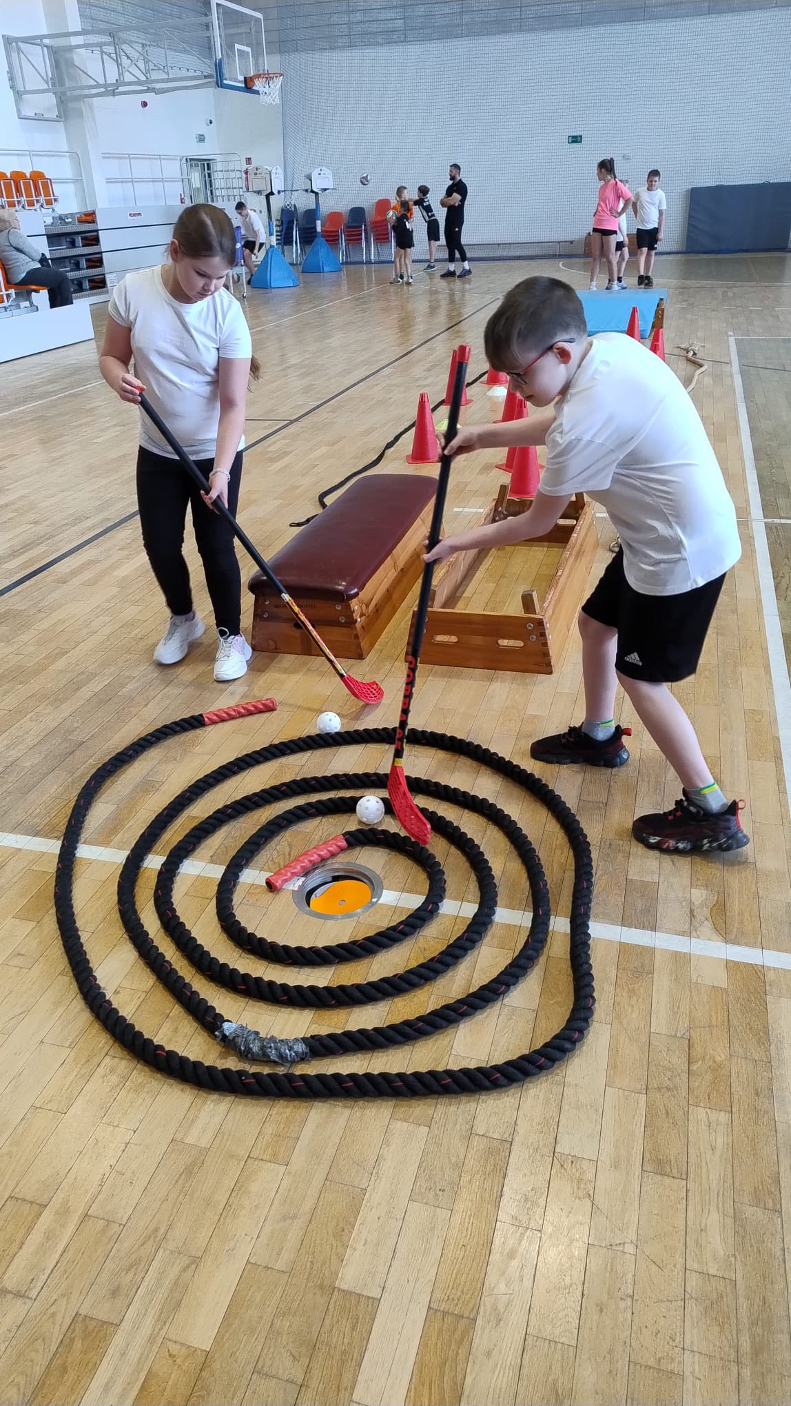 Zdjęcie przedstawia salę gimnastyczną, w której odbywają się zajęcia z unihokeja. Dwoje dzieci ćwiczy, używając kijów z czerwonymi końcówkami i kierując białe, perforowane piłki w stronę wyznaczonego celu. Cel otoczony jest grubą, czarną liną ułożoną w okrąg na drewnianej podłodze. W tle widać inne osoby oraz sprzęt sportowy i pachołki.