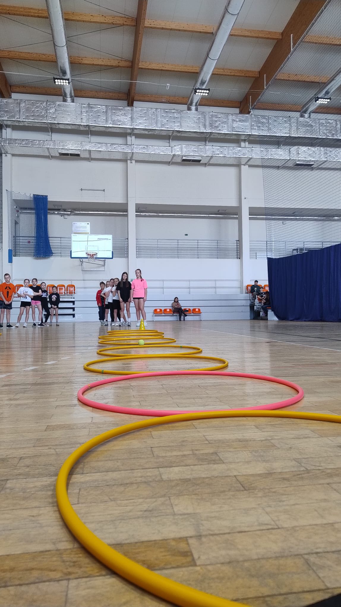 Zdjęcie przedstawia salę gimnastyczną z drewnianą podłogą, na której ułożono w linii kolorowe obręcze, naprzemiennie żółte i różowe. W tle przy ścianie stoi grupa dzieci. Na ścianie zamontowany jest kosz do koszykówki oraz niebieskie siatki zabezpieczające. Pod sufitem widoczne są drewniane belki, metalowe przewody wentylacyjne oraz oświetlenie.