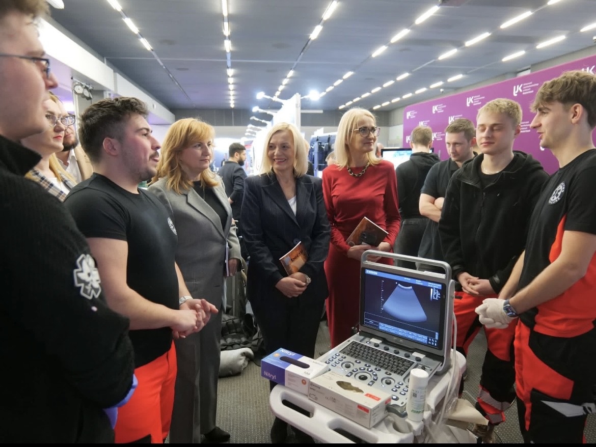 Obraz przedstawia grupę osób zgromadzonych wokół aparatu USG w jasnym wnętrzu z banerami w tle. Na ekranie urządzenia widoczny jest obraz w odcieniach szarości. Uczestnicy rozmawiają i skupiają uwagę na aparacie, co sugeruje pokaz lub prezentację sprzętu medycznego.