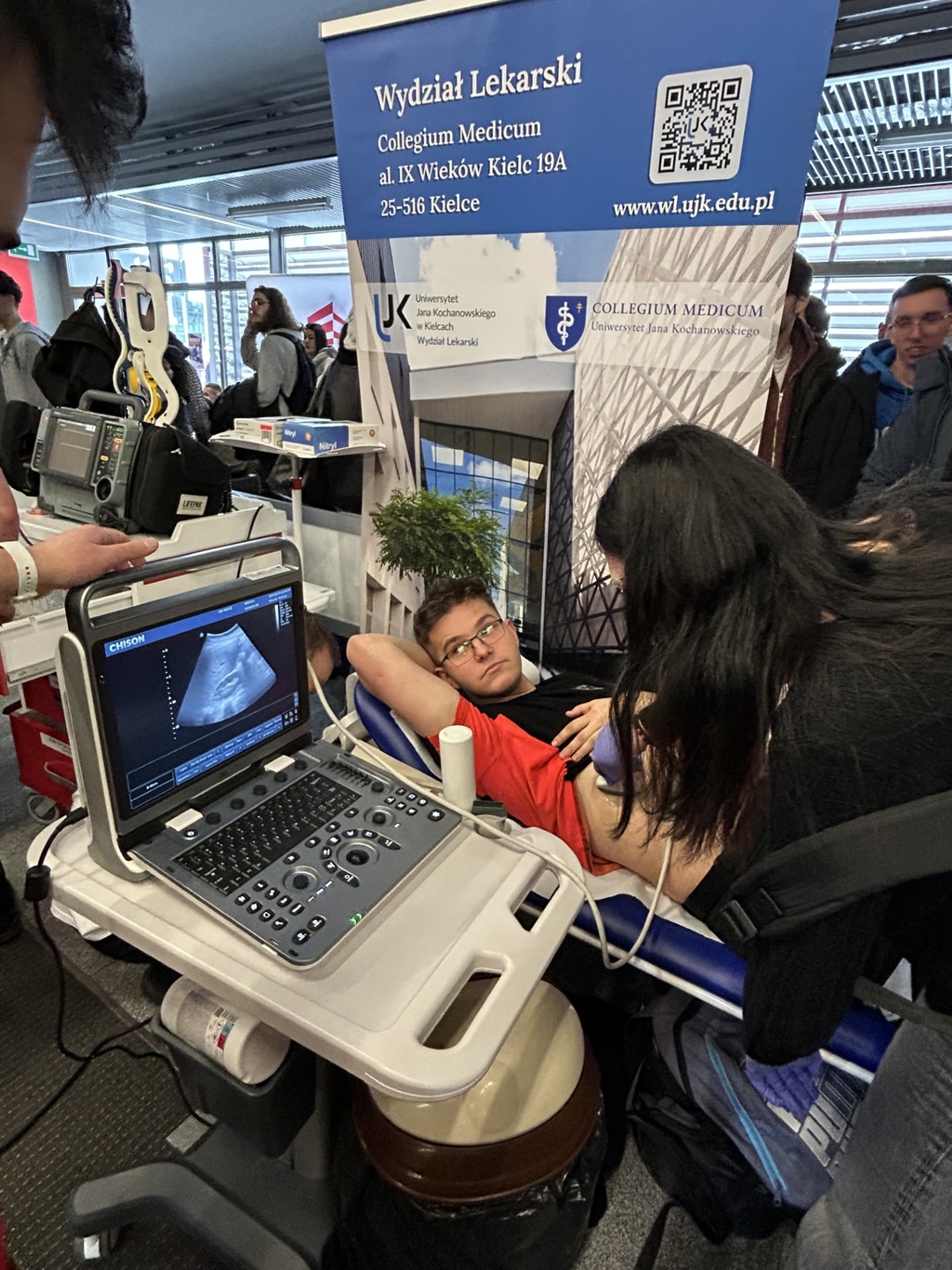 Obraz przedstawia pokaz medyczny odbywający się w ruchliwym wnętrzu. Na pierwszym planie mężczyzna leży na łóżku medycznym, a wykonywane jest u niego badanie USG. Aparatura znajduje się na wózku, a na monitorze widoczny jest obraz z badania. W tle widać grupę osób oraz baner informacyjny dotyczący wydziału medycznego, co sugeruje wydarzenie publiczne lub wystawę.