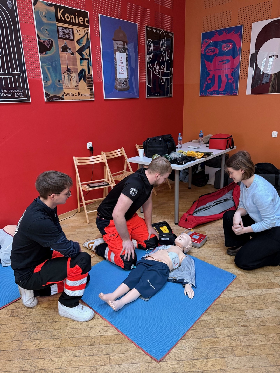 Zdjęcie przedstawia szkolenie z resuscytacji krążeniowo-oddechowej z użyciem fantomu. Zajęcia odbywają się w sali z kolorowymi ścianami i plakatami. Fantom leży na matach, a uczestnicy klęczą obok, ćwicząc i obserwując przebieg szkolenia. W pobliżu znajduje się sprzęt medyczny.