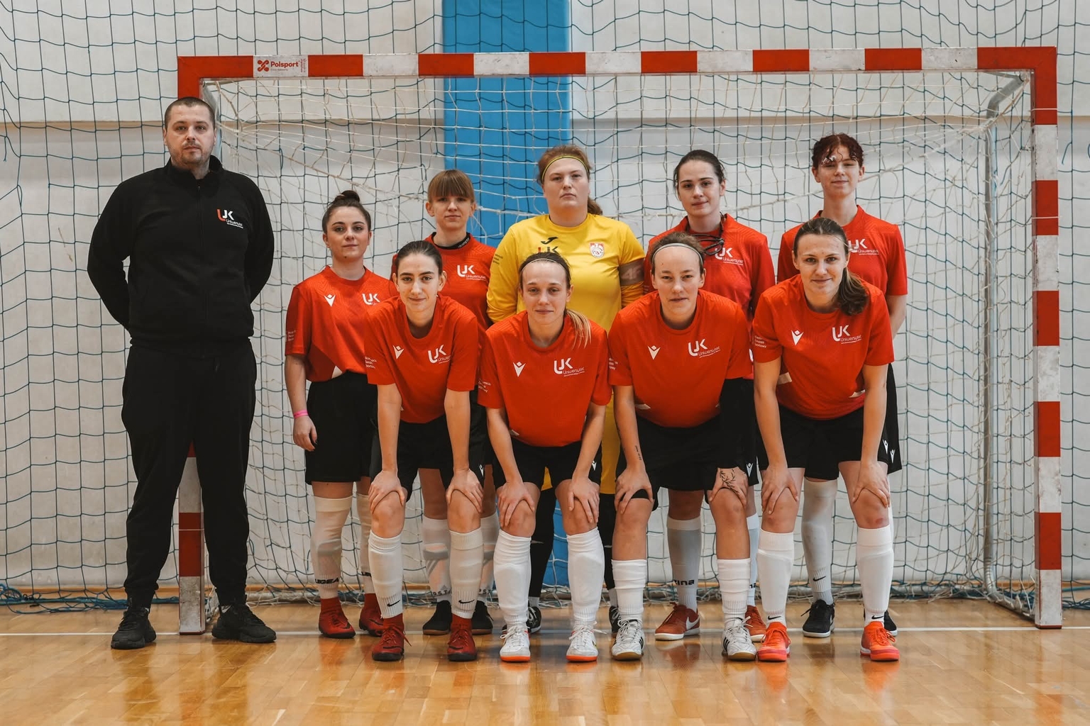 Na zdjęciu widać kobiecą drużynę sportową pozującą przed bramką w hali sportowej. Zawodniczki mają na sobie jednolite stroje sportowe, a jedna z nich wyróżnia się inną koszulką, co wskazuje na rolę bramkarki. Obok stoi trenerka. Zdjęcie wykonano w hali z drewnianą podłogą, przed bramką z siatką.