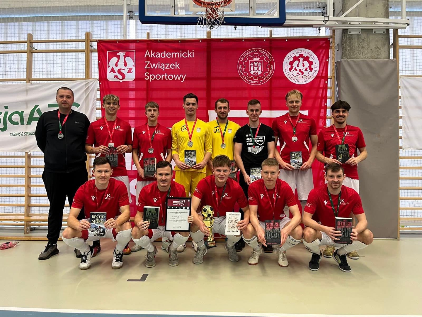 Na zdjęciu widać grupę młodych mężczyzn, prawdopodobnie drużynę sportową, pozującą w hali przed banerami. Część osób stoi, a część klęczy. Trzymają książki lub certyfikaty. Zdjęcie wykonano w hali sportowej, co wskazuje kosz do koszykówki i linie na podłodze.