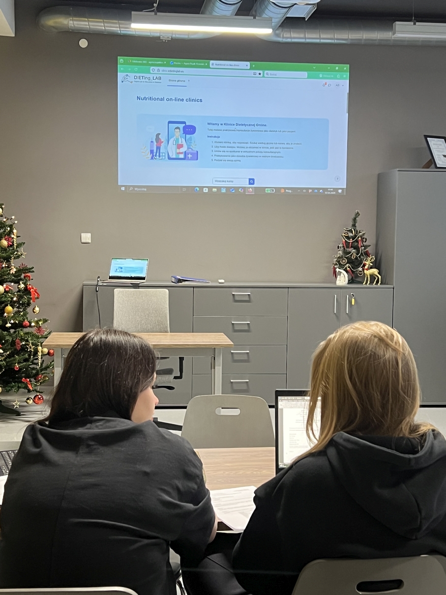 Na obrazie widać salę wykładową lub konferencyjną z rzutnikiem, na którym wyświetlana jest strona internetowa „DIETing.LAB” dotycząca internetowych poradni żywieniowych. Przed ekranem znajduje się stół z laptopem, a uczestnicy skupiają się na prezentowanej treści. W pomieszczeniu stoją także świąteczne dekoracje, w tym choinka. Wnętrze ma nowoczesny charakter.