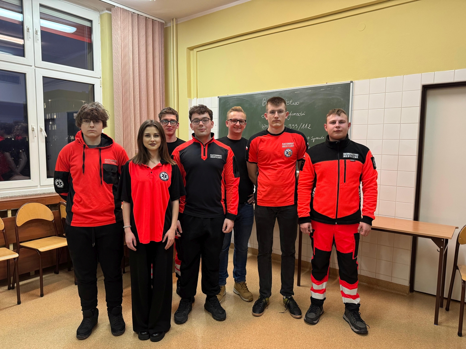 Na obrazie widać siedem osób stojących w sali przypominającej klasę, przed zieloną tablicą. W pomieszczeniu znajdują się tablica z napisami, okno z zasłonami, kilka krzeseł i stół. Atmosfera jest uporządkowana i swobodna.