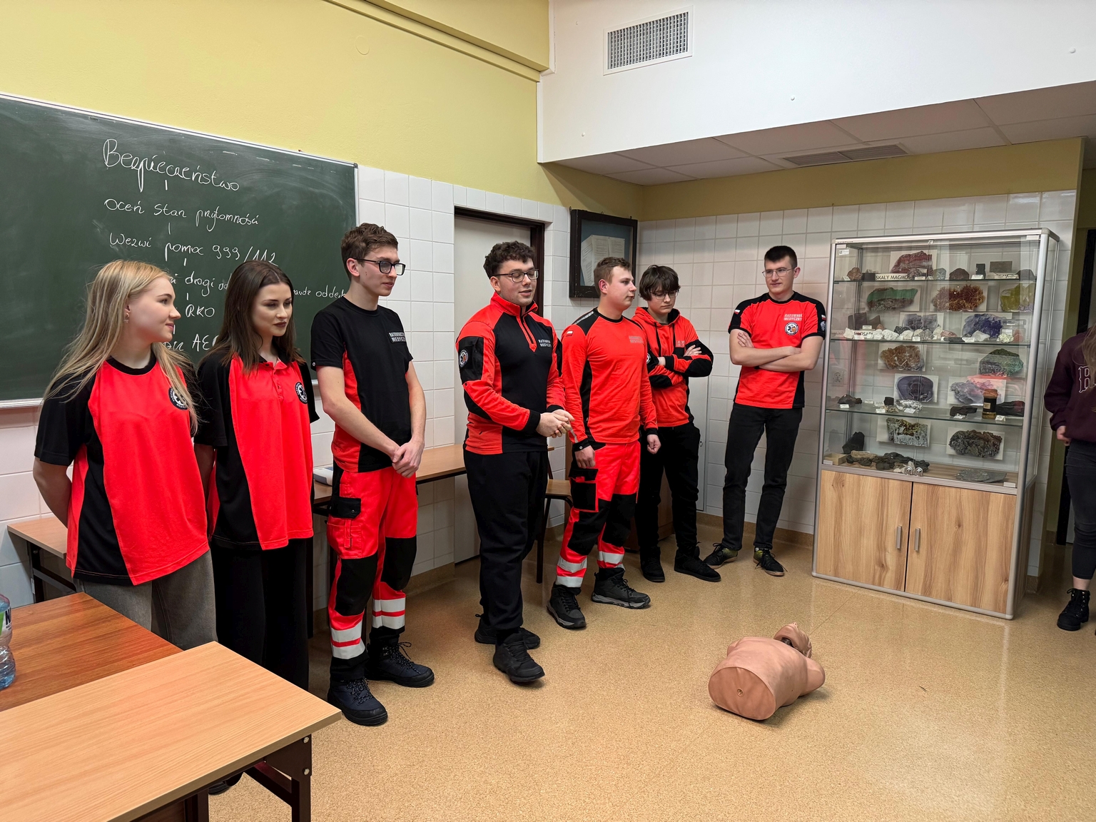 Na obrazie widać sześć osób stojących w klasie w linii, przed nimi leży manekin do ćwiczeń medycznych. W pomieszczeniu znajduje się zielona tablica z napisami, szafka ekspozycyjna z okazami skał i minerałów, beżowe ściany i jasna podłoga.