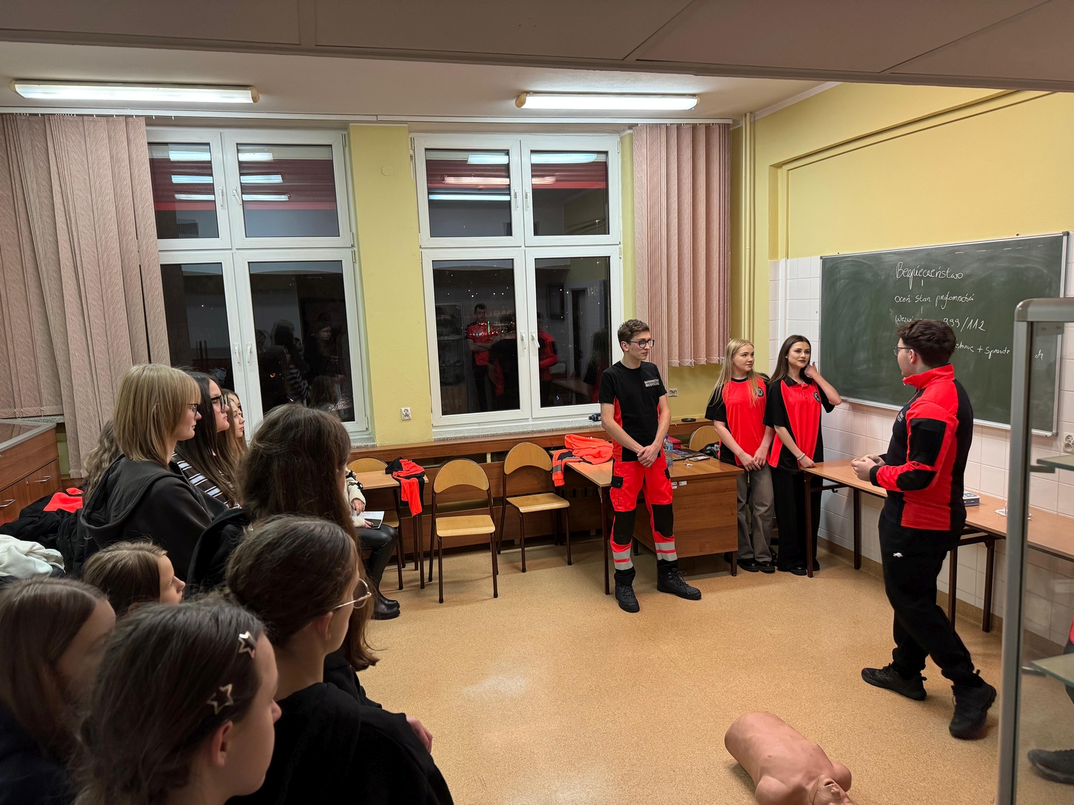 Na obrazie widać jasne wnętrze, prawdopodobnie salę lekcyjną lub szkoleniową. Grupa osób stoi w półokręgu, skupiona na osobie prowadzącej demonstrację przy tablicy. Obok niej leży manekin, sugerując szkolenie z zakresu pierwszej pomocy. W sali znajdują się duże okna z zasłonami, tablica i biurka.