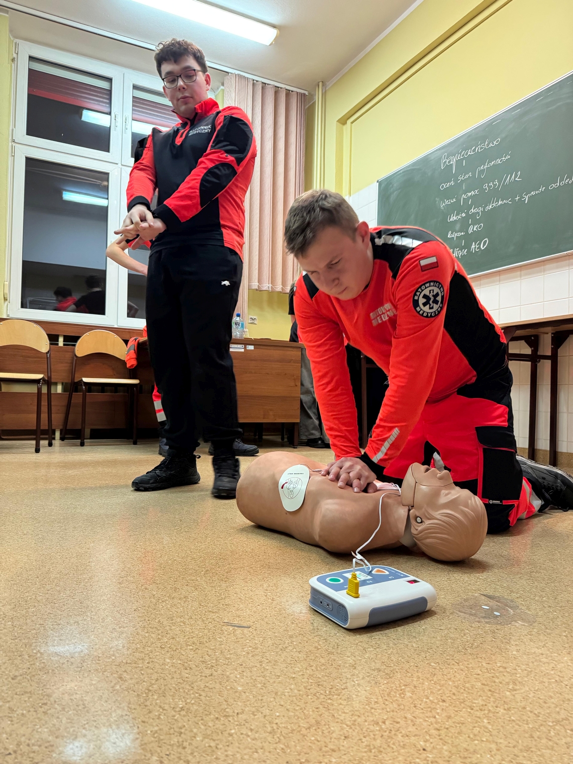 Na obrazie widać dwie osoby w sali lekcyjnej. Jedna wykonuje resuscytację na manekinie, obok znajduje się defibrylator AED. W tle jest tablica na żółtej ścianie, kilka krzeseł oraz duże okno z zasłonami. Pomieszczenie jest dobrze oświetlone.