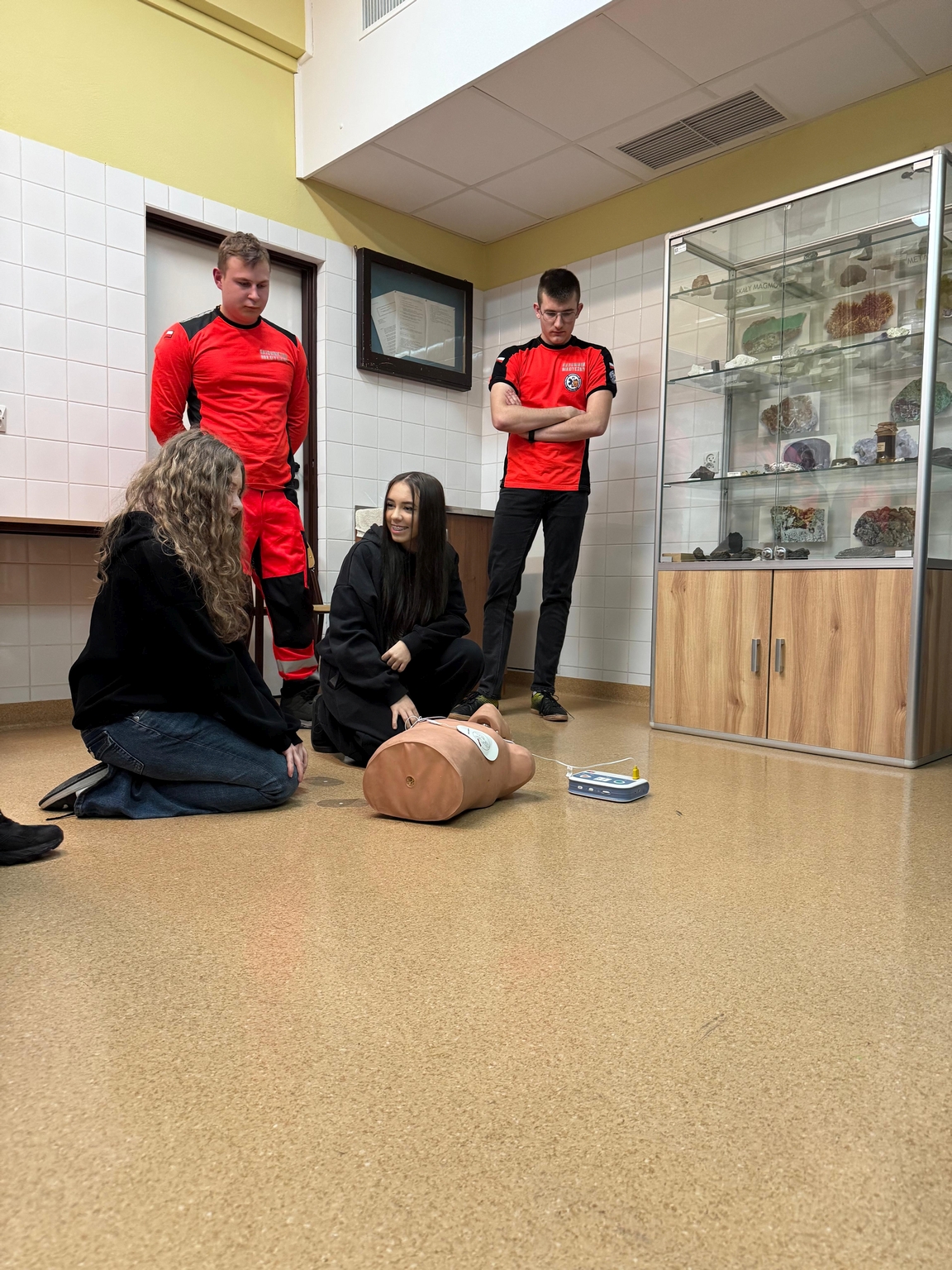 Na obrazie widać grupę czterech osób w sali przypominającej klasę z wyłożonymi kafelkami ścianami i szafką ekspozycyjną z okazami geologicznymi. Dwie osoby stoją, a dwie inne siedzą na podłodze, obsługując manekin do resuscytacji i defibrylator. Podłoga jest jasnobrązowa i gładka.