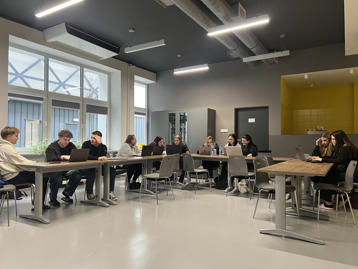 Obraz przedstawia salę dydaktyczną z kilkoma stołami, przy których używane są laptopy. Pomieszczenie jest dobrze oświetlone dzięki dużym oknom po jednej stronie. Wnętrze ma nowoczesny wygląd – jasna podłoga kontrastuje z ciemnym sufitem z widoczną instalacją oraz liniowym oświetleniem. Ściany utrzymane są w odcieniach szarości i żółci, a atmosfera sprzyja pracy i współpracy.