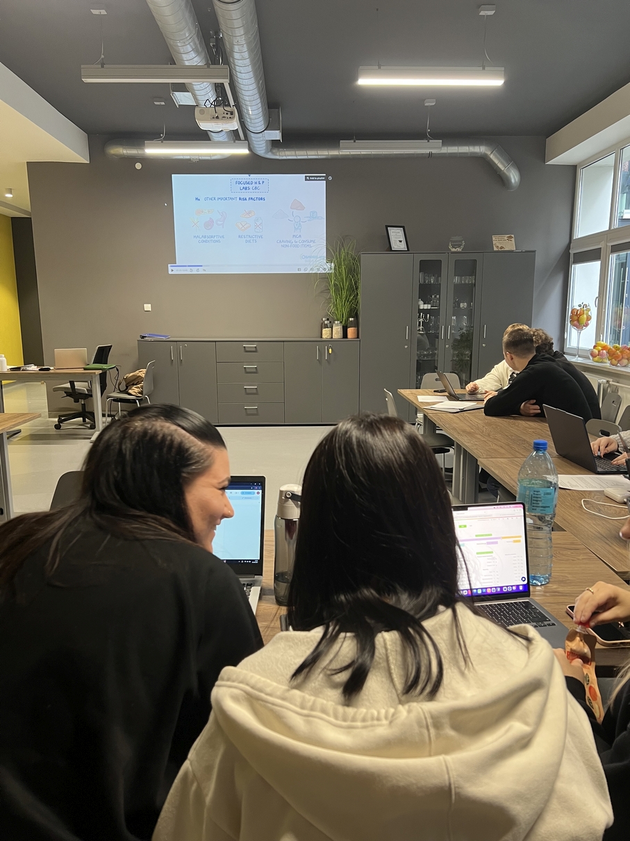 Na obrazie widać nowoczesną salę dydaktyczną lub konferencyjną, w której przy stołach używane są laptopy. Część osób pracuje wspólnie, inne indywidualnie. Na ścianie wyświetlana jest prezentacja z projektora, a pod nią znajdują się metalowe szafki. Wnętrze ma industrialny charakter z widoczną instalacją sufitową i jest dobrze doświetlone światłem dziennym wpadającym przez duże okna.