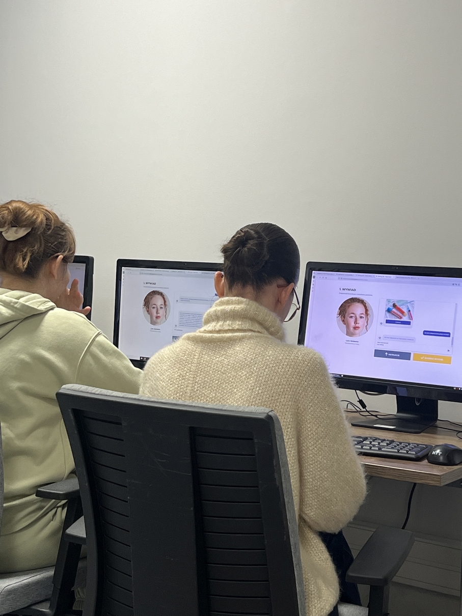 Na obrazie widać stanowisko pracy z dwoma komputerami stacjonarnymi ustawionymi na jednym biurku. Osoby siedzą obok siebie i pracują przy monitorach, na których wyświetlane są podobne strony internetowe z centralnie umieszczonym wizerunkiem oraz tekstem. Na biurku znajdują się klawiatury i myszki, a tło stanowi jasna, neutralna ściana.